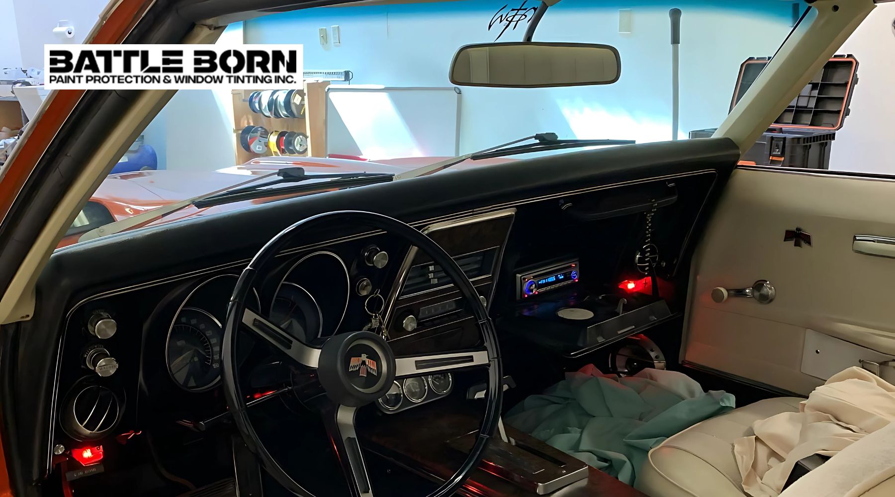 Interior of a classic car showcasing a black dashboard, chrome-accented steering wheel with an emblem, and a modern radio system installed. The door panel features a vintage handle and an emblem, while the seats are covered with cloth. The "Battle Born Paint Protection & Window Tinting Inc." logo is visible in the top left corner.