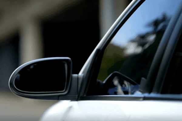 A close-up of a car's tinted side window and side mirror reflecting the surroundings, emphasizing privacy and security. The dark tint helps obscure the vehicle's interior from outside view while maintaining a sleek appearance.