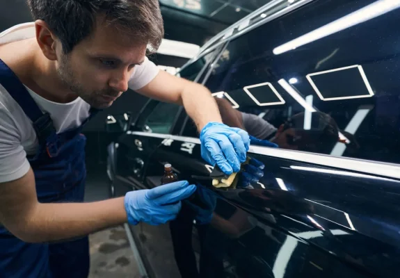 A man wearing blue gloves and overalls carefully applies a ceramic coating to the side of a black car using a small applicator pad and a bottle of coating solution. The car's glossy surface reflects the bright overhead lights in the workshop.