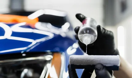 A person wearing black gloves is pouring a clear liquid from a small bottle onto a white applicator pad, preparing to apply a coating to a blue car with a shiny, reflective surface. The car's hood features bright reflections from overhead lights, and an orange cloth is visible in the background.