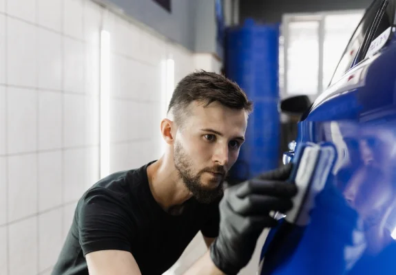 A man wearing black gloves carefully applies a protective coating or polish to the side of a shiny blue car using an applicator pad. The setting appears to be an auto detailing shop with tiled walls and bright overhead lighting.